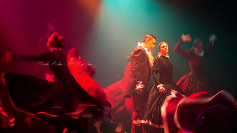 Um par com trajes de estilo vitoriano, nítido, no meio de bailarinas de saias vermelhas que giram desfocadas à sua volta.