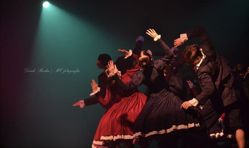 Um grupo de bailarinos com trajes de época escuros ergue as mãos ao mesmo tempo sob um único foco.
