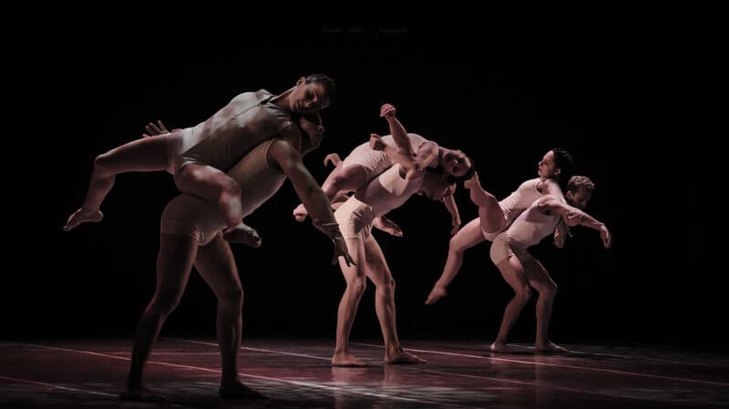 Três pares de bailarinos contemporâneos elevam-se e carregam-se uns aos outros, com figurinos cor de pele, sobre fundo preto.