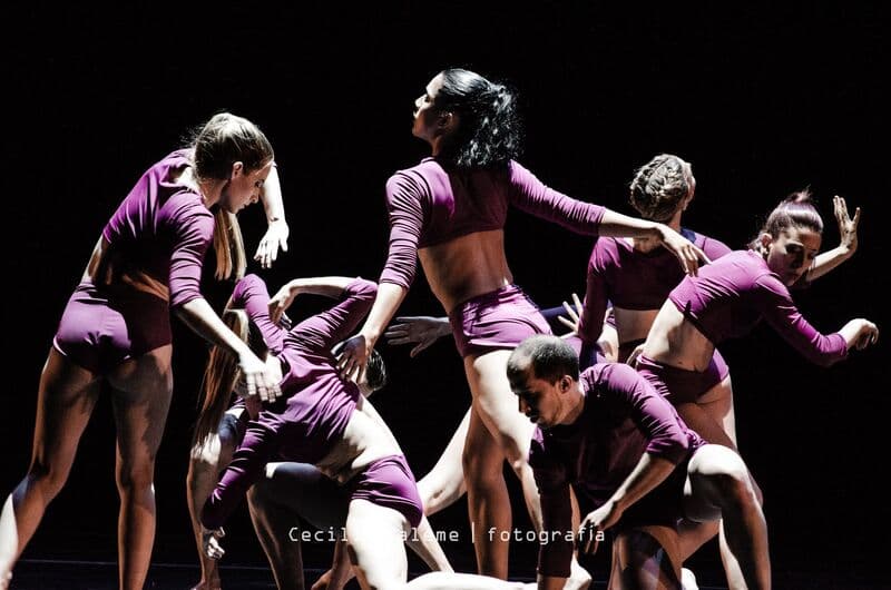 Um grupo compacto de bailarinos com fatos magenta estica-se e torce-se sob luz de palco quente.