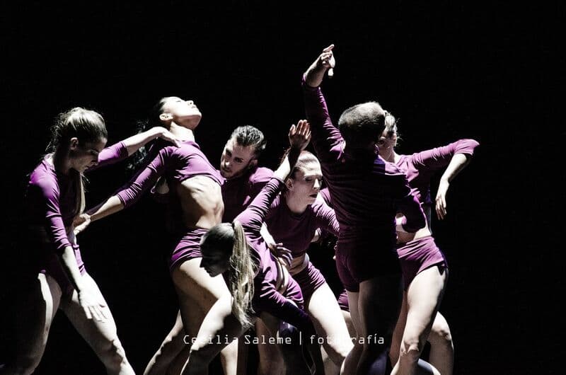 Bailarinos com fatos magenta arqueiam-se e esticam-se num grupo compacto; uma mulher, de rosto iluminado, inclina-se para trás.