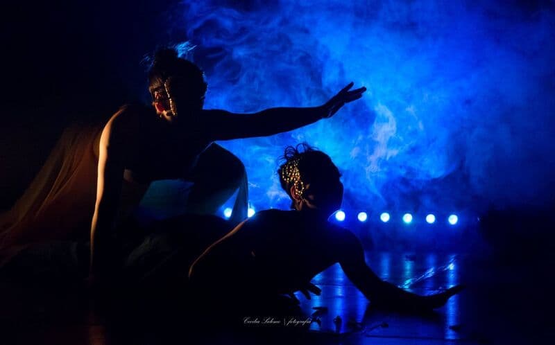 Dois bailarinos mascarados, em silhueta, iluminados por fumo azul e uma fila de luzes de palco atrás.