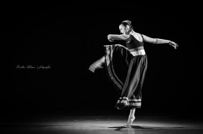 Uma bailarina a solo de saia escura gira em pontas sob um único foco sobre fundo preto, a preto e branco.