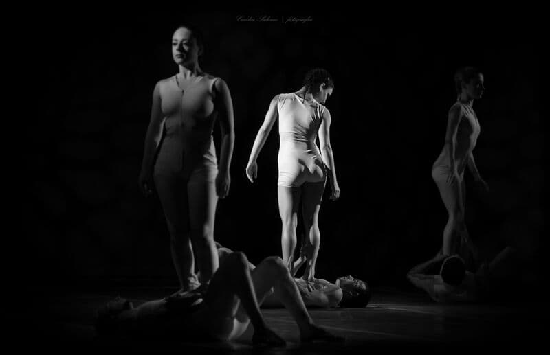 Bailarinos de pé equilibram-se sobre os pares deitados num palco escuro, a preto e branco.