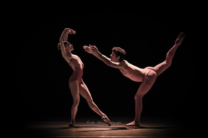 Um par de ballet vestido de cor-de-rosa dança um pas de deux, ele com a perna estendida bem alto para trás, sobre fundo preto.