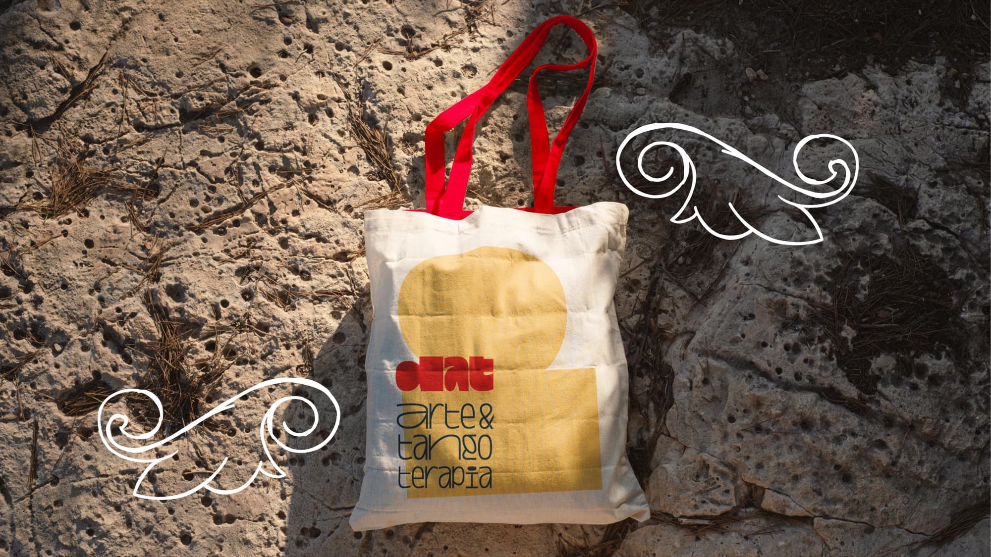 Una bolsa de tela color crudo con asas rojas y el logo de DAT sobre una forma amarilla, apoyada en una piedra, con adornos dibujados a mano alrededor.