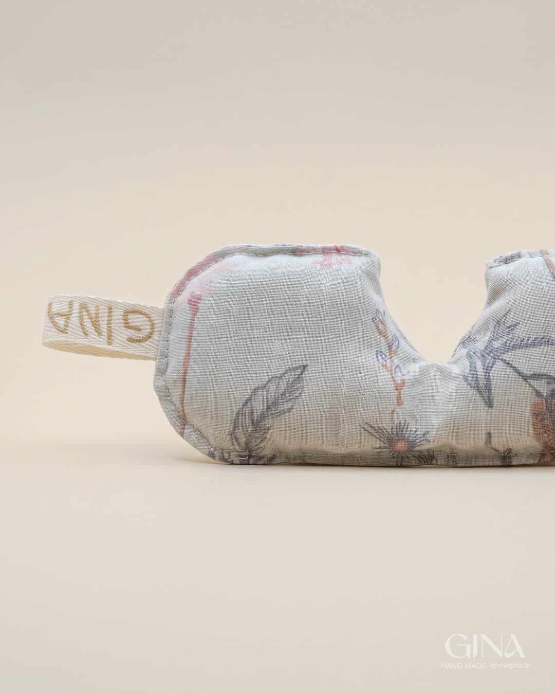 A GINA eye pillow in cream floral linen with the fabric tag, on a beige background.
